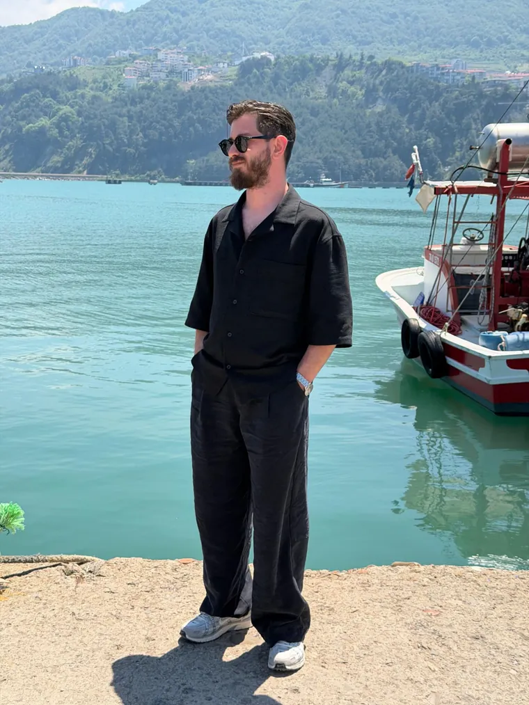 Murat Akkaş, siyah keten gömlek ve pantolonla Karadeniz'de bir limanda; yanında kırmızı beyaz bir balıkçı teknesi