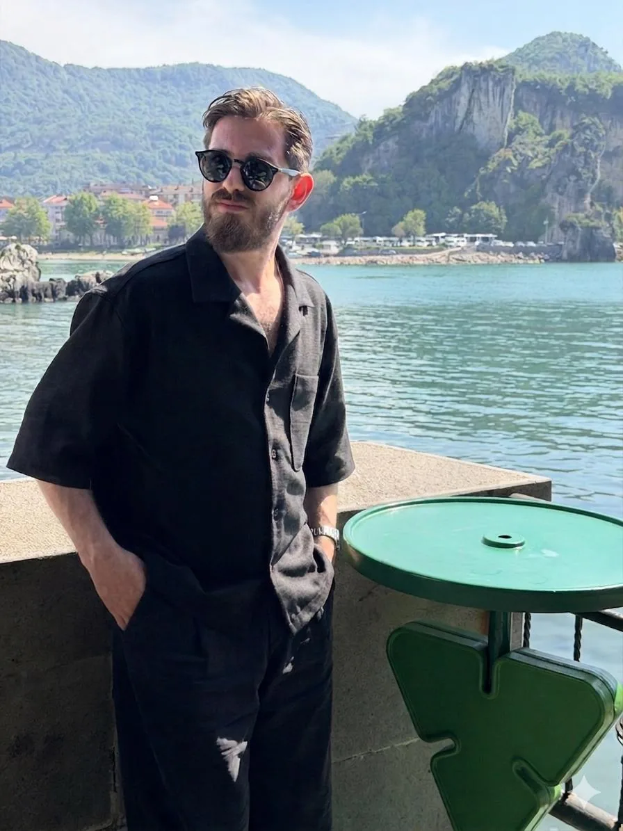 Murat Akkaş, güneş gözlüğüyle deniz kenarındaki taş duvara yaslanmış; arkada yeşil tepeler ve kayalıklar